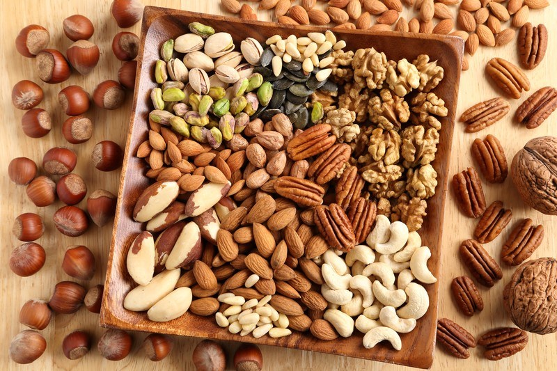 A square wooden dish filled with a variety of nuts is surrounded by several types of loose nuts on a wooden table.