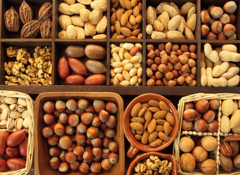 This photo shows an overhead view of several types of nuts, ranging from chestnuts and almonds to pecans and walnuts, in wooden compartments and baskets.