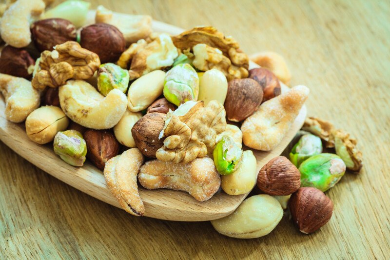 This photo shows a wooden scoop filled with several types of nuts that are spilling out of it onto a wooden table.