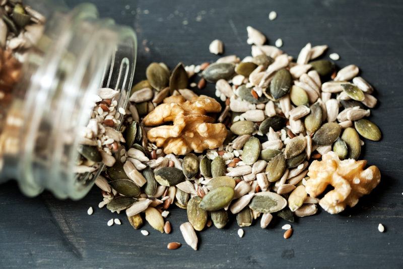 Various nuts and seeds spilling onto the table from a jar. Pumpkin seeds and sesame seeds are the most common