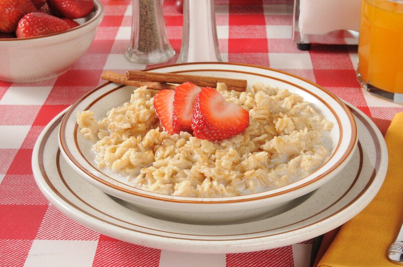 This photo shows a bowl of oatmeal with milk and strawberries on a red and white tablecloth.