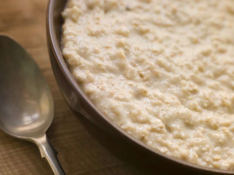 This photo shows a closeup of a bowl of cooked oatmeal near a spoon on a wooden table.