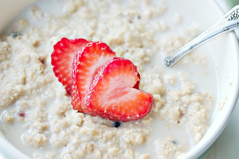 This photo shows an overhead view of a white bowl of oatmeal with milk and strawberry with a silver spoon.