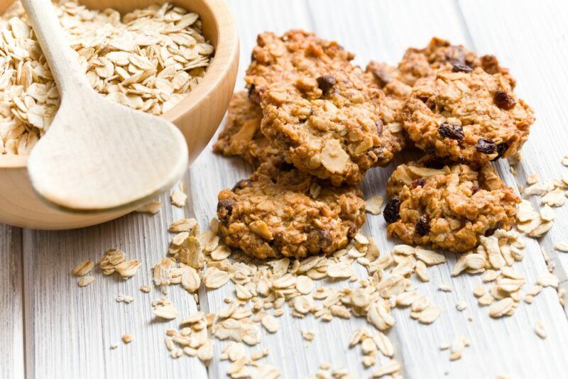 A wooden table with oatmeal cookies, plus a bowl of oats and more oats scattered on the table