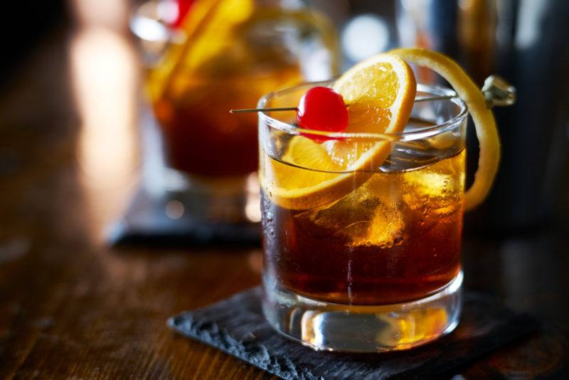 Two old fashioned cocktails with citrus slices and a glace cherry. One is in focus, while the other is not.