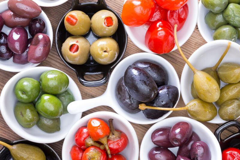 Several types of olives and pimentos rest in white bowls on a wooden surface.