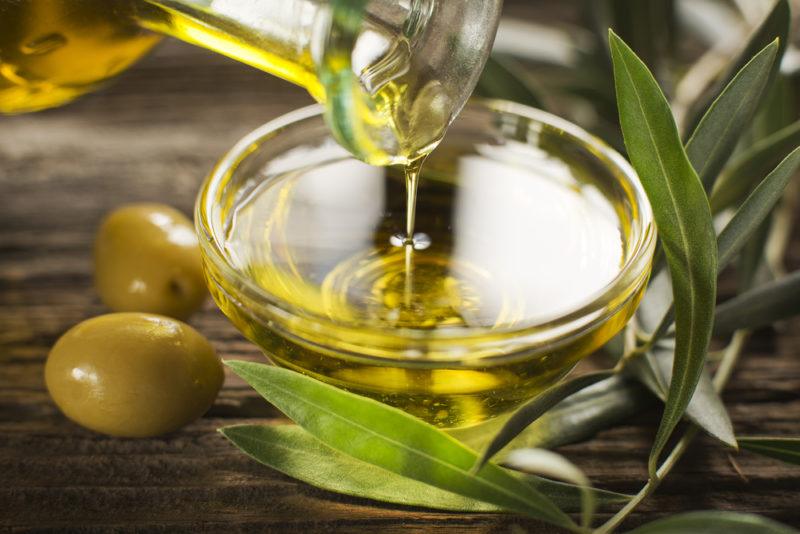 A small glass dish that is being filled with olive oil from a jug, next to some olive leaves and two green olives