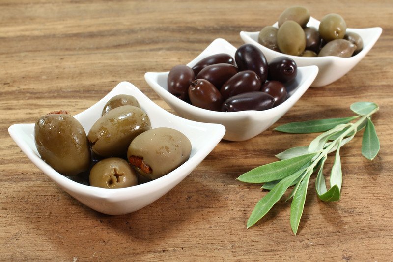 Three square white bowls filled with green and black olives rest on a wooden table next to green leaves.