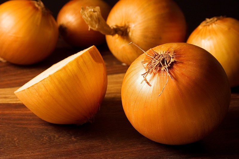 This photo shows five whole yellow onions and one half yellow onion lying on a wooden table.