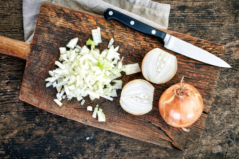 This photo shows an overhead view of a cutting board with a knife, a whole yellow onion, a halved yellow onion, and some chopped yellow onion, overlaying a white kitchen towel on a wooden table.