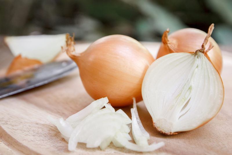 Two whole raw onions on a cutting board and one that has been cut in half, with pieces of onion and a knife too