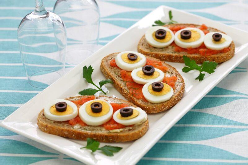 A white rectangular plate with three open faced sandwiches that use fish, eggs, and olives as ingredients