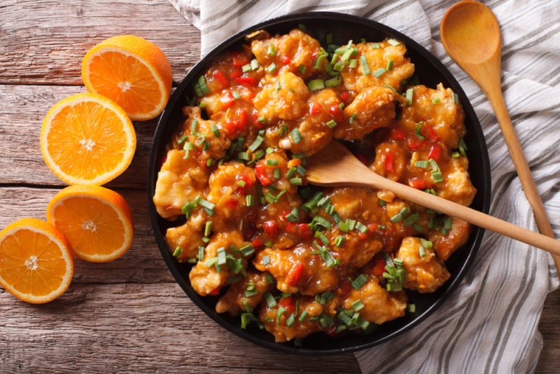 A black frypan with orange Chinese chicken next to sliced oranges