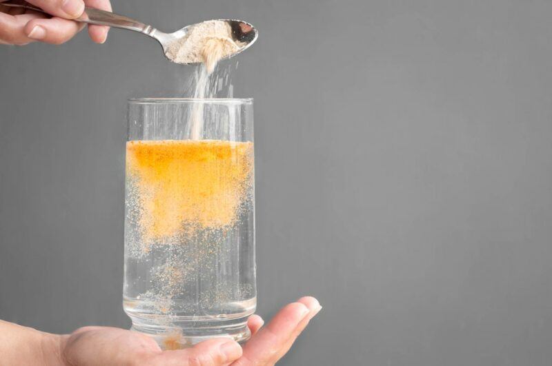 A hand holding a glass of water, where a spoon of Metamucil is being poured in and turns yellow in the water