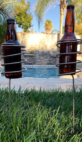 Two beer holders with beer staked into the grass in front of a pool. 