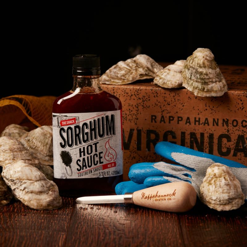 Rappahannock Oyster Co box in the back with oysters scattered around and on the box and table.  Also featured is a bottle of Sorghum hot sauce ,  Rappahannock  oyster shucking knife, and shucking gloves