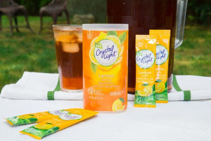 A container of Crystal Light tea powder, with individual sachets and a glass of the prepared ice tea in the background