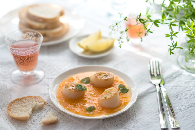 A white plate with a tomato-based sauce and three scallops, next to a plate of lemon wedges, bread, and two glasses of rose