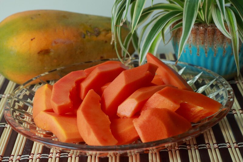 This photo shows papaya chunks in a glass bowl near an uncut papaya and a spider plant on a bamboo mat.