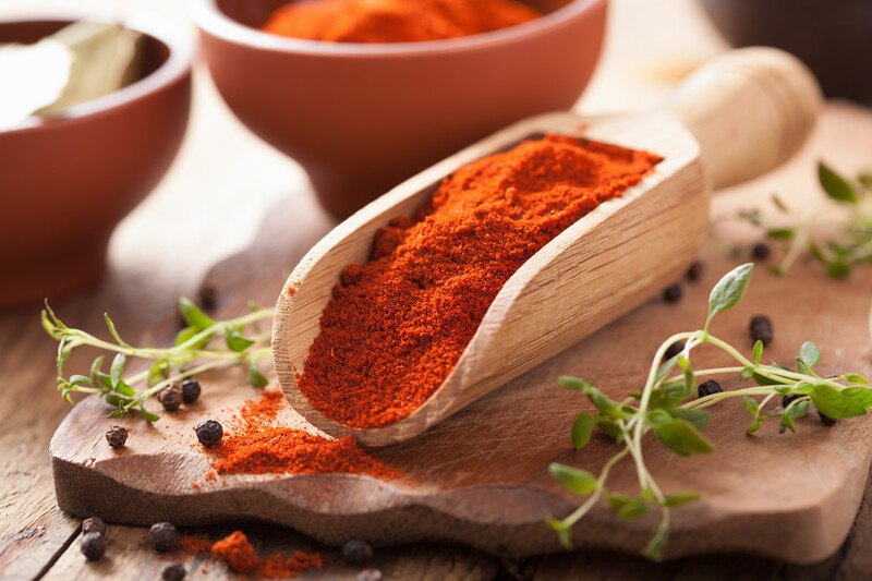 a wooden scoop full of paprika resting on a wooden chopping board with fresh herbs and loose black peppercorns around it, with a couple of wooden bowls at the back