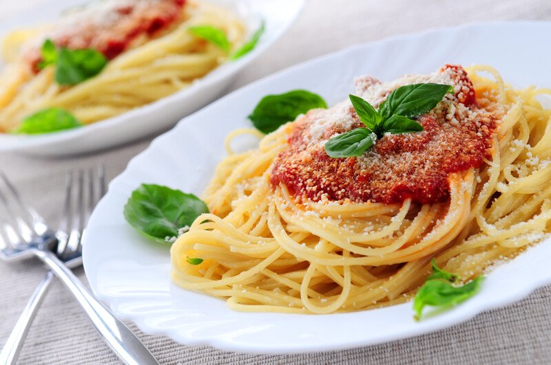 Two white plates with spaghetti, marinara sauce, and green leaf garnishes rest on a beige tablecloth next to two silver forks.