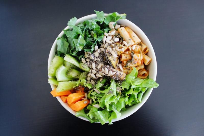 A white bowl with pasta salad and various greens