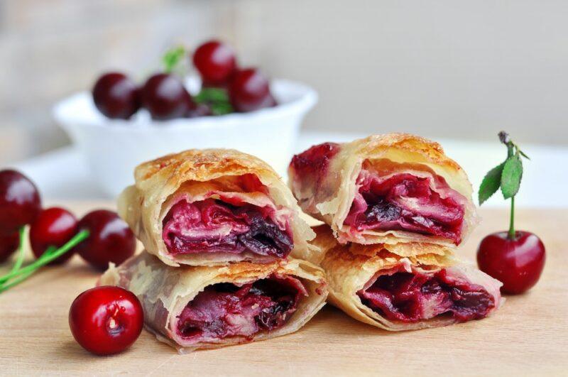 Four pastries made using strawberries or cherries, with a bowl of cherries in the background