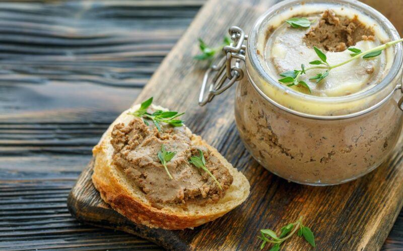 A glass jar of liver pate next to a piece of bread with more of the same pate