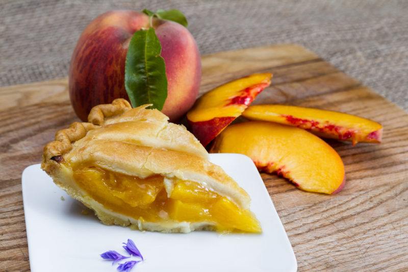 A wooden board with a full peach, some peach slices, and a piece of peach pie