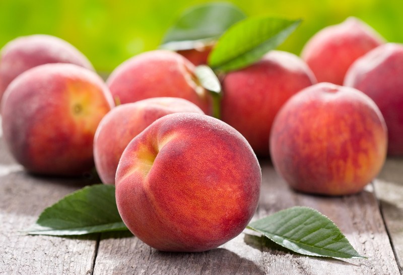 12 red-colored peaches with green leaves rest on a wooden surface.