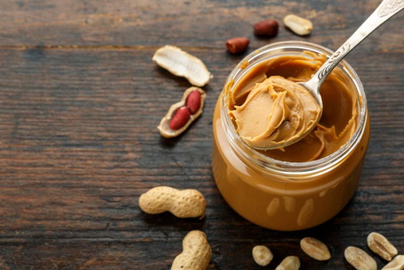 A jar containing peanut butter with a spoon and various peanuts scattered around