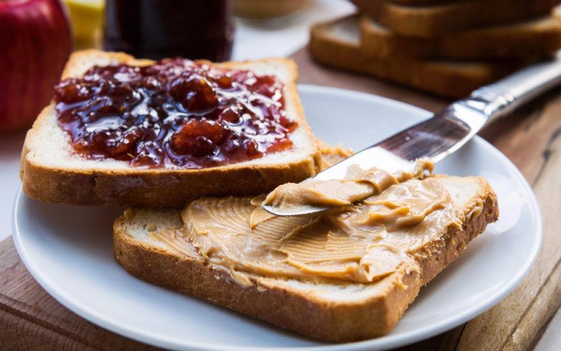 A piece of bread or toast with peanut butter and a knife, next to another with jam