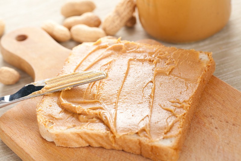 Peanut butter on white bread sitting on a wooden cutting board, with a butter knife leaning against the bread.  A jar of peanut butter and shelled peanuts are in the background