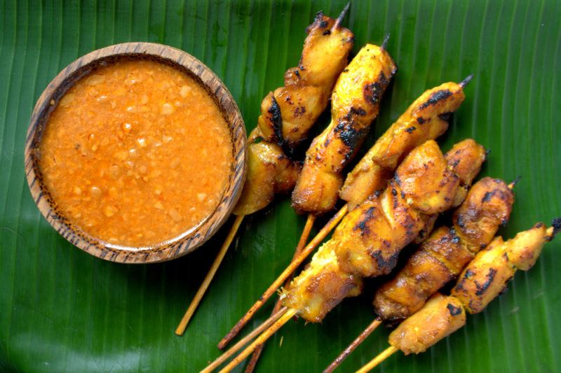 Half a dozen or so kebabs with satay chicken next to peanut sauce on a large green leaf