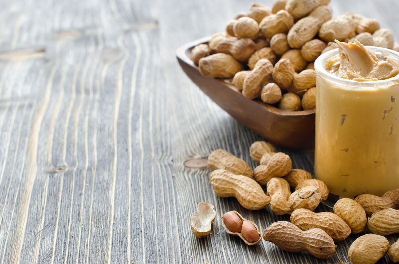 A brown bowl of peanuts, with peanuts on a table and a jar of peanut butter