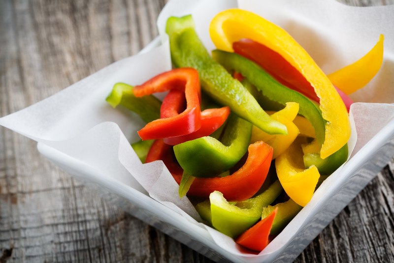 This photo shows a white square bowl with several green, red, and yellow pepper strips in it, on top of a wooden table.