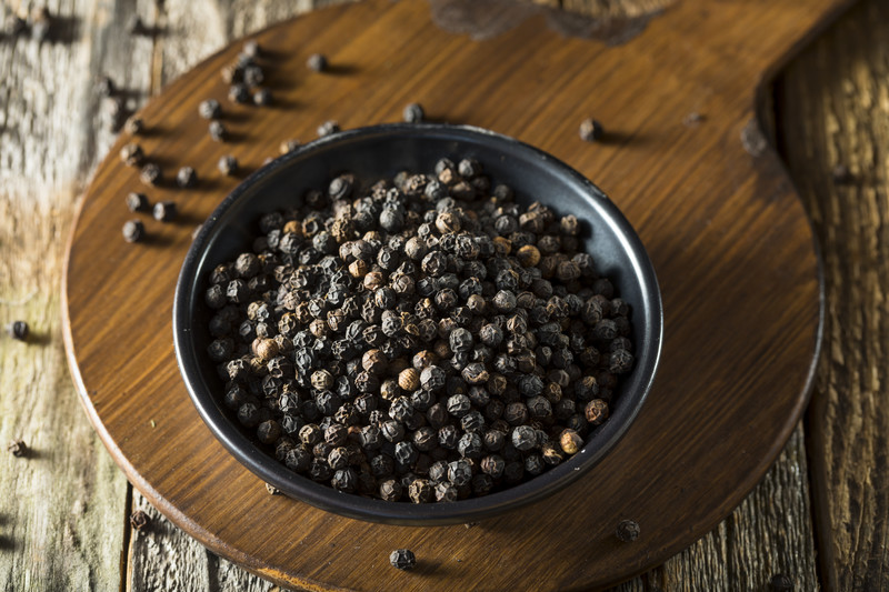 Black ceramic bowl full of peppercorns resting on a wood palette on top of an old wooden surface.