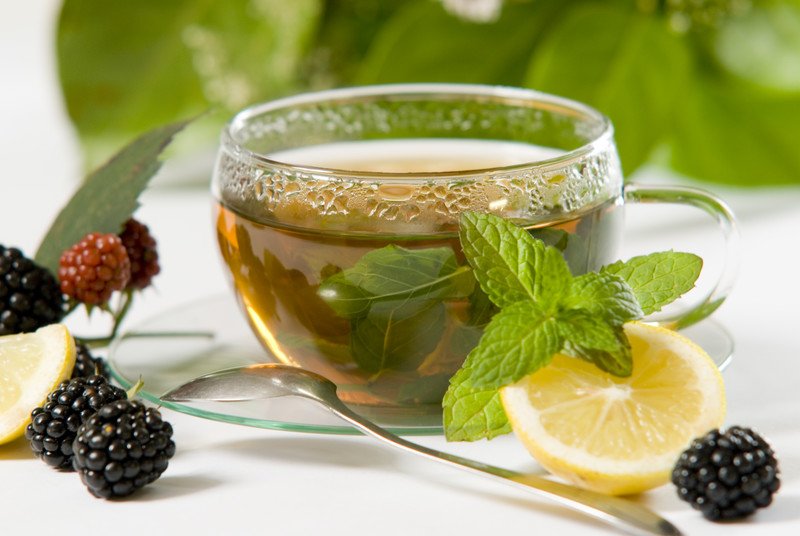 This photo shows a cup of peppermint tea near a sprig of peppermint, a lemon slice, and a few blackberries on a white tablecloth.