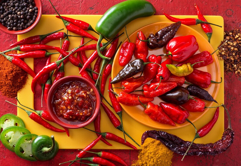 This photo shows an overhead view of a yellow cutting board topped with and surrounded by several types of hot peppers in red and green, as well as a bowl of pepper sauce, a bowl of peppercorns, and three piles of pepper flakes and powders on a red tabletop.