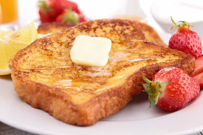 A white plate containing a slice of well cooked French toast with butter, next to strawberries