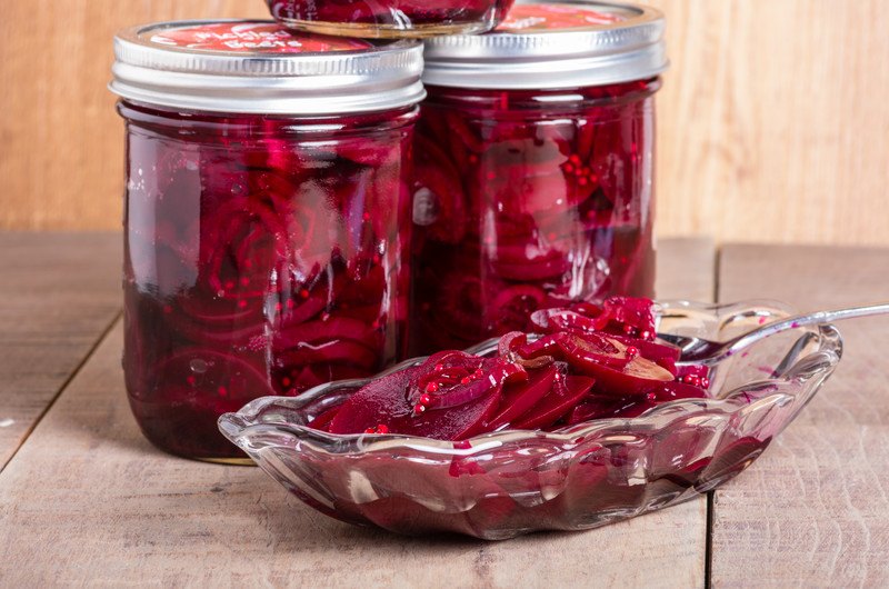 This photo shows a few jars of pickled beets near a glass dish filled with pickled beets and a spoon on a wooden table.