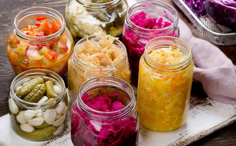 Several open jars of pickled foods, including pickled cucumbers, purple cabbage, and kimchi, rest on a rustic white painted board.