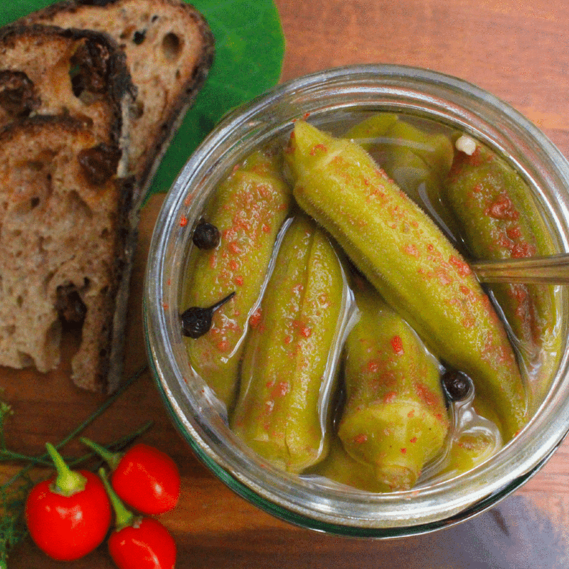 Pickled okra in an open jar with cayenne sprinkled on top, next to the jar three red cherry peppers, and in the upper left corner are two slices of raisin bread.