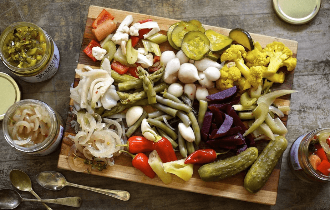 Wooden cutting board with an array of pickled vegetables like onions, asparagus, mini peppers, garlic, pearl onion, cauliflower, cucumbers, beets, and green beans.  3 jars of pickles surrounding the cutting board and three silver spoons in the lower left corner 