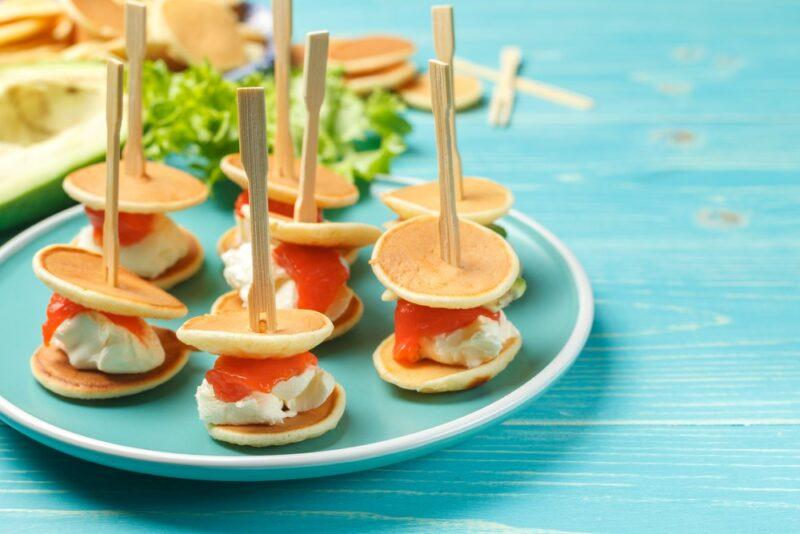 A light blue table and a light blue plate containing picnic sandwiches that have been made with cream cheese, salmon, and mini pancakes, with a stick through them