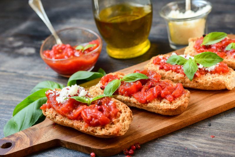 A wooden board with bruschetta, with fresh tomatoes and basi, next to a jar of sauce, olives, and cheese