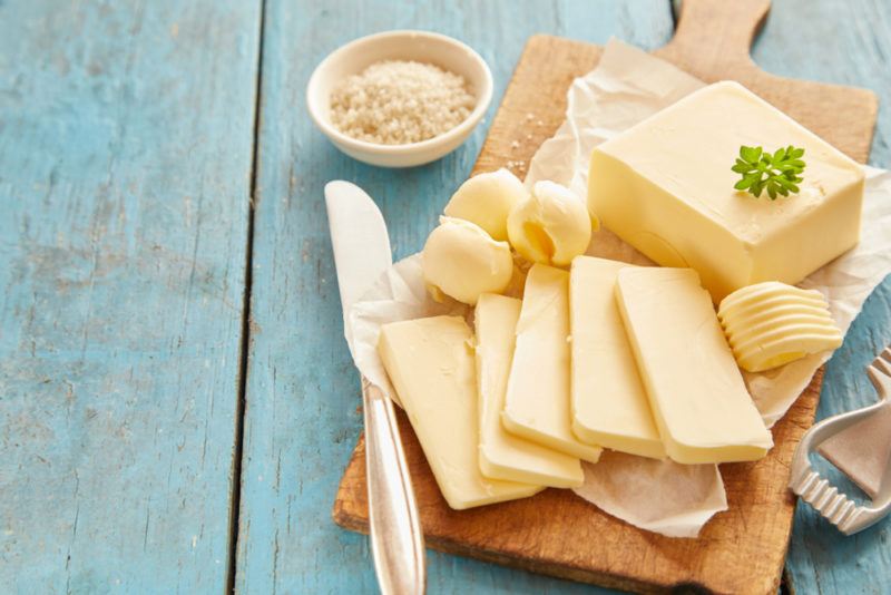 A light blue table with a wooden board that contains butter which has been sliced and there are some curls of it too, next to a knife