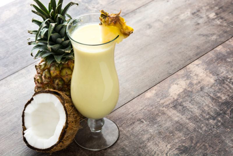 A tall glass containing a pina colada next to a pineapple and a coconut