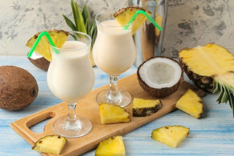 A wooden board with two pina colada cocktails and pineapple wedges, with pineapple and coconut on the table