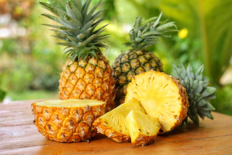 Two full pineapples, a pineapple cut into two halves, and two pineapple wedges rest on a wooden surface.